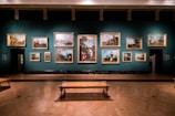 A dimly lit art gallery displays an array of classical paintings, each framed in ornate gold, hung against a teal wall. The floor features polished wood, and there is a simple wooden bench in the foreground. The paintings depict various historical or architectural scenes.