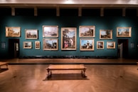 A dimly lit art gallery displays an array of classical paintings, each framed in ornate gold, hung against a teal wall. The floor features polished wood, and there is a simple wooden bench in the foreground. The paintings depict various historical or architectural scenes.