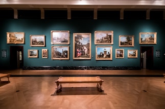 A dimly lit art gallery displays an array of classical paintings, each framed in ornate gold, hung against a teal wall. The floor features polished wood, and there is a simple wooden bench in the foreground. The paintings depict various historical or architectural scenes.