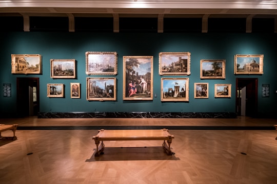A dimly lit art gallery displays an array of classical paintings, each framed in ornate gold, hung against a teal wall. The floor features polished wood, and there is a simple wooden bench in the foreground. The paintings depict various historical or architectural scenes.