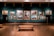 A dimly lit art gallery displays an array of classical paintings, each framed in ornate gold, hung against a teal wall. The floor features polished wood, and there is a simple wooden bench in the foreground. The paintings depict various historical or architectural scenes.