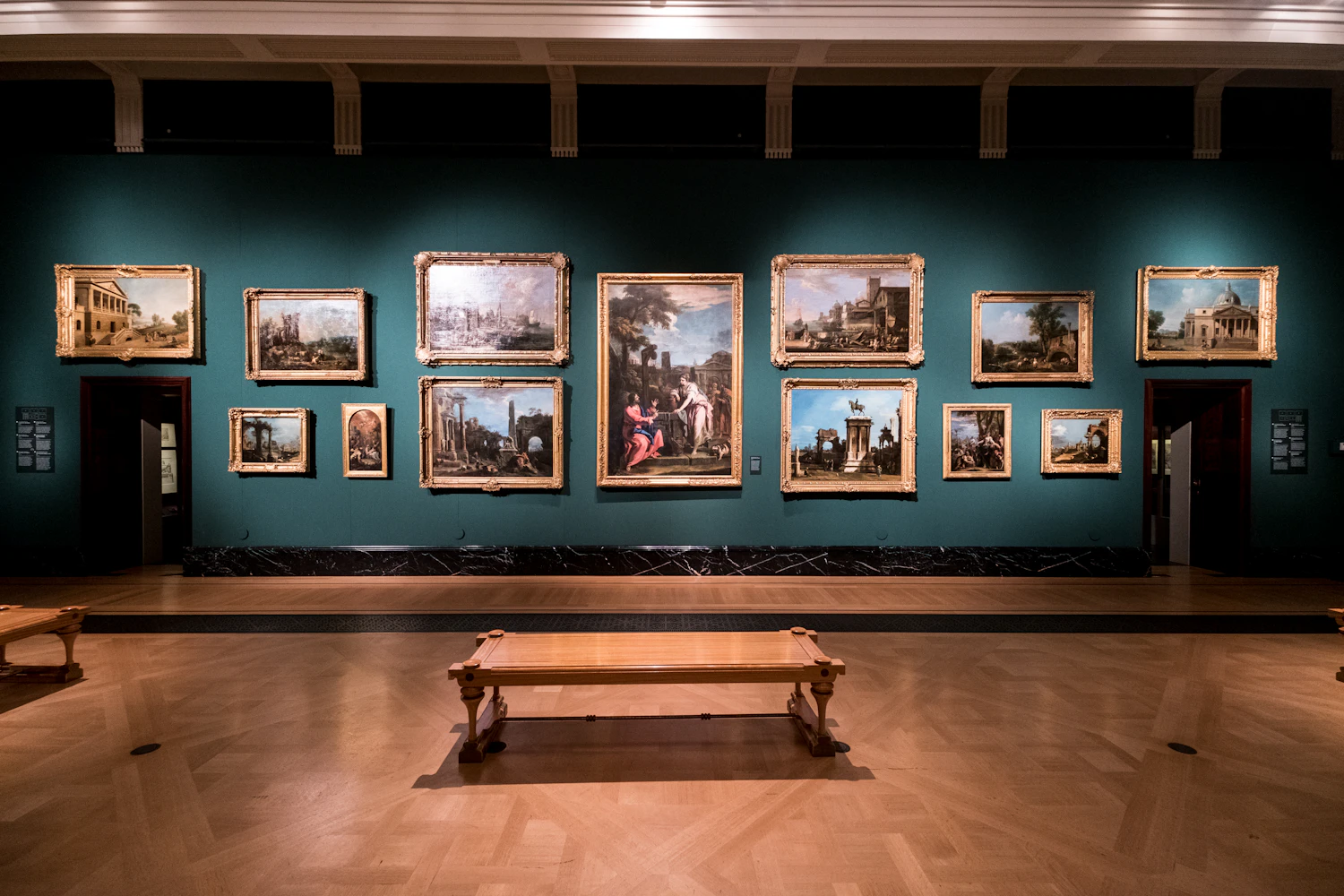 The finished hang: a deep green wall carrying nineteen framed works in three tiers above a bench