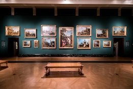 A dimly lit art gallery displays an array of classical paintings, each framed in ornate gold, hung against a teal wall. The floor features polished wood, and there is a simple wooden bench in the foreground. The paintings depict various historical or architectural scenes.