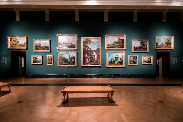 A dimly lit art gallery displays an array of classical paintings, each framed in ornate gold, hung against a teal wall. The floor features polished wood, and there is a simple wooden bench in the foreground. The paintings depict various historical or architectural scenes.
