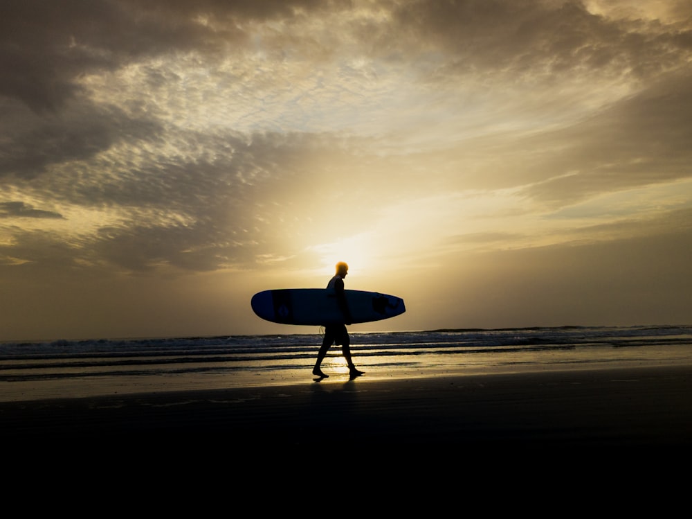 man holding surfboard walking on beach during sunset