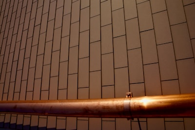 A section of a building facade with a pattern of rectangular tiles arranged in a linear grid. A copper-colored metallic pipe runs horizontally across the lower part of the image, with a smooth surface and metallic sheen contrasted against the matte tiles.