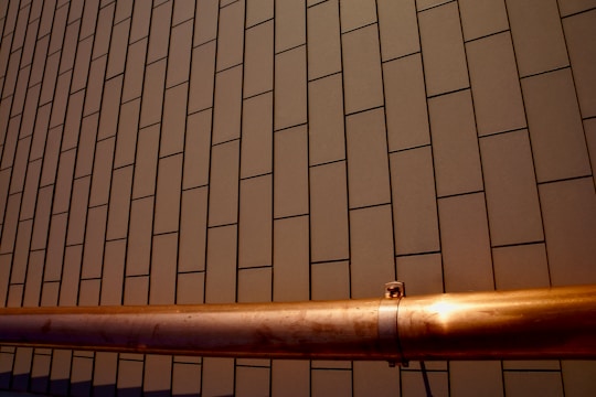 A section of a building facade with a pattern of rectangular tiles arranged in a linear grid. A copper-colored metallic pipe runs horizontally across the lower part of the image, with a smooth surface and metallic sheen contrasted against the matte tiles.