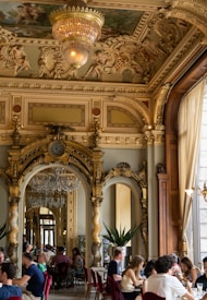 An opulent, ornately decorated interior featuring intricate gold moldings and a large chandelier hanging from a richly adorned ceiling. The room has grand arches and mirrors that reflect the elegant design, with people sitting at tables in a luxurious setting.