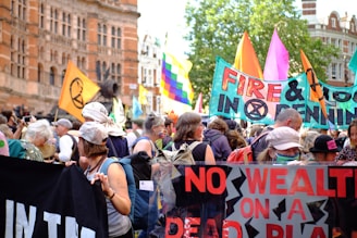 A large group of people gathered for a protest, holding colorful banners and signs. The banners contain messages related to climate change, displaying slogans like 'No Wealth On A Dead Planet' and 'Fire & Flood in Openings'. The crowd is diverse, with individuals wearing different types of clothing, hats, and masks. The background features historical buildings with detailed architecture.