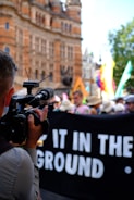 A person is holding and using a video camera to film a group of people involved in a public demonstration or protest. A black banner with partially visible white text is being held up by the protesters. The background shows an ornate historical building and some trees.