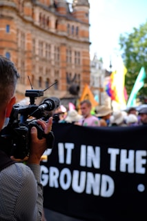 A person is holding and using a video camera to film a group of people involved in a public demonstration or protest. A black banner with partially visible white text is being held up by the protesters. The background shows an ornate historical building and some trees.