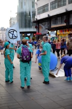 A group of people dressed in green and blue scrubs gather in a busy urban area. One person is holding a flag with a black symbol on it. They appear to be engaged in some kind of demonstration or public event as onlookers pass by. There are also street shops and a modern building in the background.