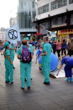 A group of people dressed in green and blue scrubs gather in a busy urban area. One person is holding a flag with a black symbol on it. They appear to be engaged in some kind of demonstration or public event as onlookers pass by. There are also street shops and a modern building in the background.