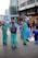 A group of people dressed in green and blue scrubs gather in a busy urban area. One person is holding a flag with a black symbol on it. They appear to be engaged in some kind of demonstration or public event as onlookers pass by. There are also street shops and a modern building in the background.
