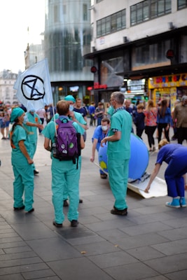 A group of people dressed in green and blue scrubs gather in a busy urban area. One person is holding a flag with a black symbol on it. They appear to be engaged in some kind of demonstration or public event as onlookers pass by. There are also street shops and a modern building in the background.