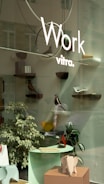 A storefront display featuring minimalist and modern decor with several small shelves mounted on a glass surface. Each shelf holds various decorative objects, including a wooden bird figure and abstract sculptures. In the foreground, there is a small table with papers and a decorative elephant sculpture. The display is complemented by a couple of indoor plants.