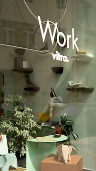 A storefront display featuring minimalist and modern decor with several small shelves mounted on a glass surface. Each shelf holds various decorative objects, including a wooden bird figure and abstract sculptures. In the foreground, there is a small table with papers and a decorative elephant sculpture. The display is complemented by a couple of indoor plants.