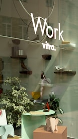 A storefront display featuring minimalist and modern decor with several small shelves mounted on a glass surface. Each shelf holds various decorative objects, including a wooden bird figure and abstract sculptures. In the foreground, there is a small table with papers and a decorative elephant sculpture. The display is complemented by a couple of indoor plants.