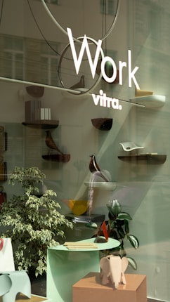 A storefront display featuring minimalist and modern decor with several small shelves mounted on a glass surface. Each shelf holds various decorative objects, including a wooden bird figure and abstract sculptures. In the foreground, there is a small table with papers and a decorative elephant sculpture. The display is complemented by a couple of indoor plants.