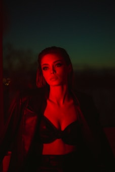 A person with dramatic red lighting casts a moody and enigmatic look. The background is dark with hints of green or blue, suggesting an evening or night setting. The person appears to be wearing a black leather jacket and a top that leaves the shoulders bare, adding to a bold and confident appearance.
