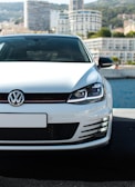 A white Volkswagen car is prominently displayed in the foreground, parked on a pavement with a waterfront and a city skyline in the background. The focus is on the front grille and headlights, with modern high-rise buildings partially visible under a clear sky.