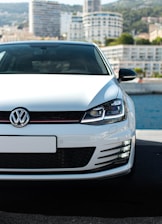 A white Volkswagen car is prominently displayed in the foreground, parked on a pavement with a waterfront and a city skyline in the background. The focus is on the front grille and headlights, with modern high-rise buildings partially visible under a clear sky.