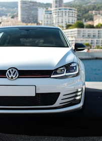 A white Volkswagen car is prominently displayed in the foreground, parked on a pavement with a waterfront and a city skyline in the background. The focus is on the front grille and headlights, with modern high-rise buildings partially visible under a clear sky.