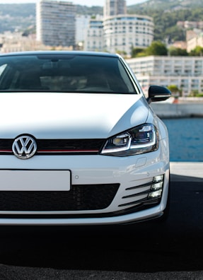 A white Volkswagen car is prominently displayed in the foreground, parked on a pavement with a waterfront and a city skyline in the background. The focus is on the front grille and headlights, with modern high-rise buildings partially visible under a clear sky.