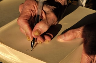 A close-up of an elderly person's hands, one holding a pen and the other steadying a notebook. The hands are weathered and aged, with noticeable fingernails and wrinkles. The setting suggests the person is writing or taking notes in a well-lit environment, with sunlight casting warm shadows.