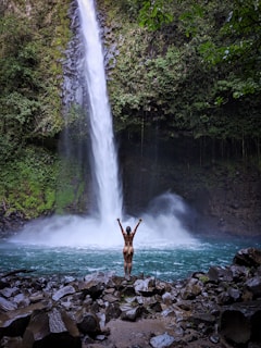 man standing on rocky shore near waterfalls during daytime