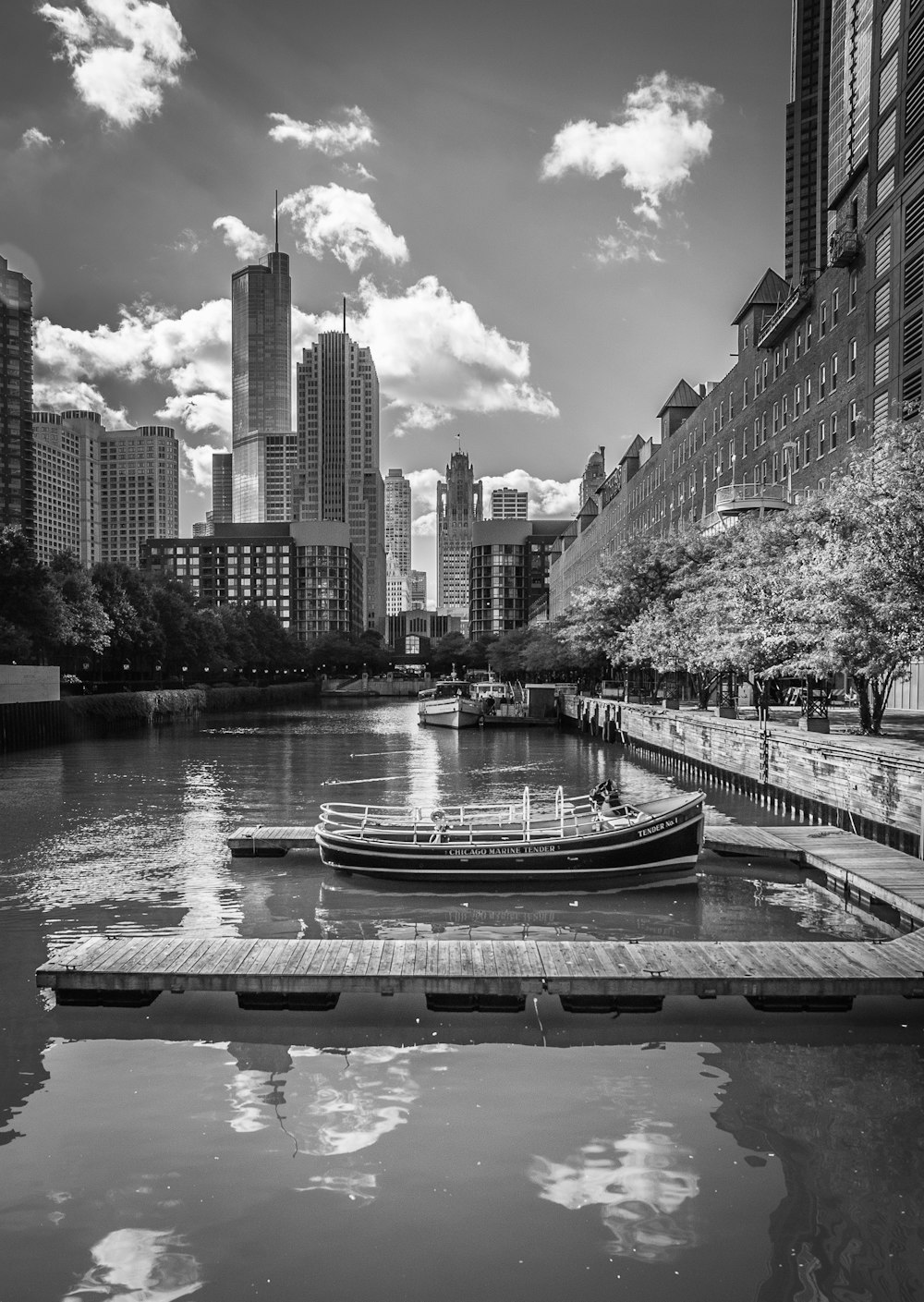 Foto en escala de grises de un barco en el río cerca de los edificios de la ciudad