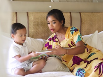 A woman and a young child are sitting together on a bed. The woman is wearing a colorful dress and is engaging with the child, who is holding a picture book. Their setting appears cozy and intimate, with neutral-toned bedding and a woven headboard.