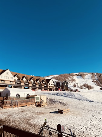 A clear blue sky above a winter resort with a row of ski lodge buildings. Snow-covered slopes in the background with ski lifts visible. Foreground displays a wooden deck with ski racks and some equipment.