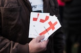 A person is holding several pamphlets with a large red cross design on them. The text on the pamphlets includes 'THE CROSS OF LOVE.' The individual is wearing a brown jacket, and the background is blurred, suggesting an outdoor environment.