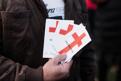 A person is holding several pamphlets with a large red cross design on them. The text on the pamphlets includes 'THE CROSS OF LOVE.' The individual is wearing a brown jacket, and the background is blurred, suggesting an outdoor environment.