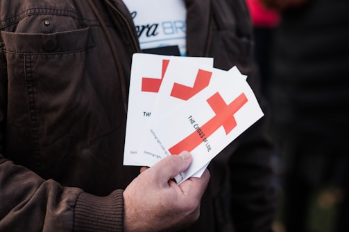 A person is holding several pamphlets with a large red cross design on them. The text on the pamphlets includes 'THE CROSS OF LOVE.' The individual is wearing a brown jacket, and the background is blurred, suggesting an outdoor environment.