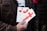 A person is holding several pamphlets with a large red cross design on them. The text on the pamphlets includes 'THE CROSS OF LOVE.' The individual is wearing a brown jacket, and the background is blurred, suggesting an outdoor environment.