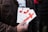 A person is holding several pamphlets with a large red cross design on them. The text on the pamphlets includes 'THE CROSS OF LOVE.' The individual is wearing a brown jacket, and the background is blurred, suggesting an outdoor environment.