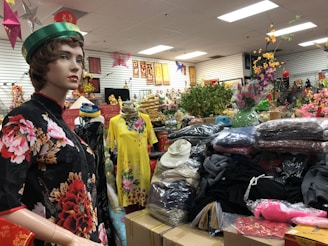 A mannequin dressed in a black floral dress is placed amid a clutter of various items inside a store. The store appears to sell traditional clothing and decorative items, with an array of colorful dresses, hats, and artificial flowers on display. There are framed art pieces on the walls and a stack of folded clothing on the shelves.