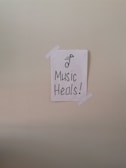 A simple piece of white paper is taped to a wall with two strips of white tape. The paper has a drawing of a musical note and the words 'Music Heals!' written in bold, black lettering.