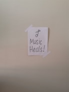 A simple piece of white paper is taped to a wall with two strips of white tape. The paper has a drawing of a musical note and the words 'Music Heals!' written in bold, black lettering.