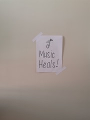 A simple piece of white paper is taped to a wall with two strips of white tape. The paper has a drawing of a musical note and the words 'Music Heals!' written in bold, black lettering.
