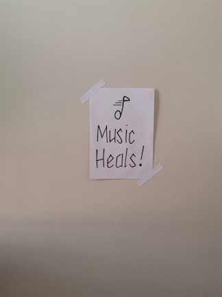 A simple piece of white paper is taped to a wall with two strips of white tape. The paper has a drawing of a musical note and the words 'Music Heals!' written in bold, black lettering.