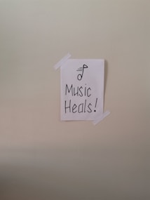 A simple piece of white paper is taped to a wall with two strips of white tape. The paper has a drawing of a musical note and the words 'Music Heals!' written in bold, black lettering.