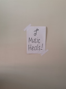 A simple piece of white paper is taped to a wall with two strips of white tape. The paper has a drawing of a musical note and the words 'Music Heals!' written in bold, black lettering.