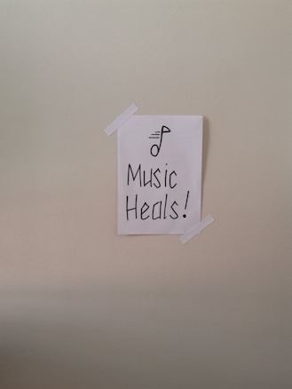 A simple piece of white paper is taped to a wall with two strips of white tape. The paper has a drawing of a musical note and the words 'Music Heals!' written in bold, black lettering.