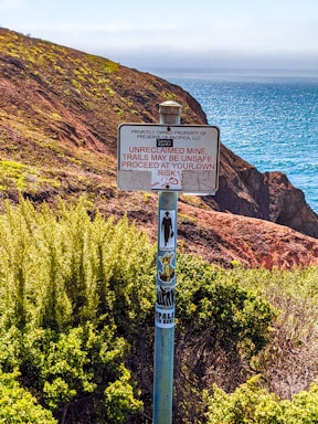 A sign is posted on a metal pole surrounded by dense greenery, with rugged coastal cliffs and the ocean visible in the background. The sign warns about an unclaimed mine and unsafe trails, advising visitors to proceed at their own risk. The pole is adorned with various stickers, including a figure silhouette.