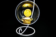 A stack of gold-colored Ethereum coins placed inside a transparent spherical container held by a white, spiral stand on a black background.
