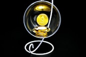 A stack of gold-colored Ethereum coins placed inside a transparent spherical container held by a white, spiral stand on a black background.
