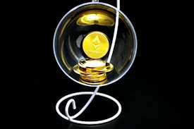 A stack of gold-colored Ethereum coins placed inside a transparent spherical container held by a white, spiral stand on a black background.
